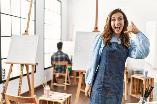 Young artist woman at art studio smiling doing phone gesture with hand and fingers like talking on the telephone. communicating concepts.