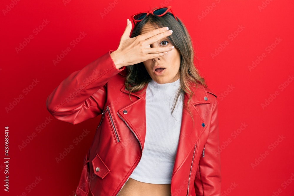 Young brunette woman wearing red leather jacket peeking in shock covering face and eyes with hand, looking through fingers with embarrassed expression. Young brunette woman wearing red leather jacket peeking in shock covering face and eyes with hand, looking through fingers with embarrassed expression.