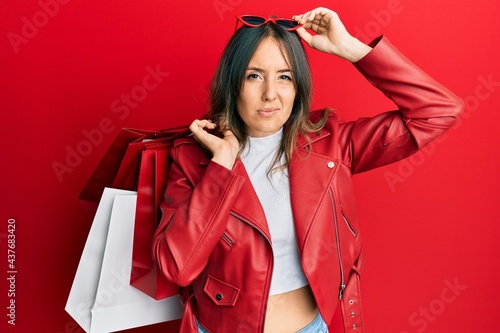 Young brunette woman holding shopping bags clueless and confused expression. doubt concept.