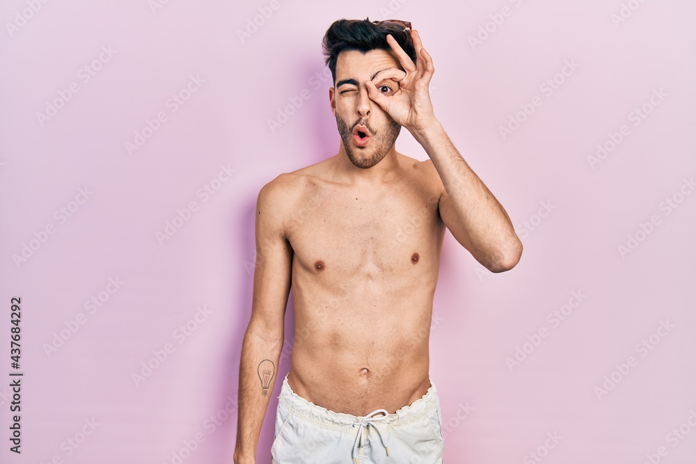Fototapeta premium Young hispanic man wearing swimwear shirtless doing ok gesture shocked with surprised face, eye looking through fingers. unbelieving expression.