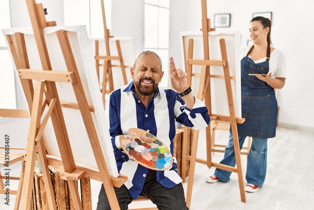 Senior artist man at art studio gesturing finger crossed smiling with hope and eyes closed. luck and superstitious concept. Senior artist man at art studio gesturing finger crossed smiling with hope and eyes closed. luck and superstitious concept.