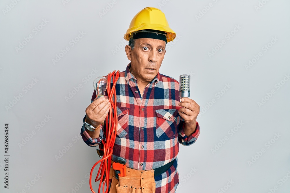 Senior hispanic man wearing handyman uniform holding led lightbulb and incandescent bulb clueless and confused expression. doubt concept.
