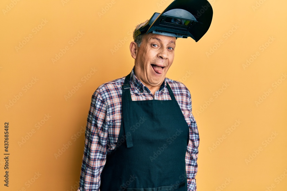 Handsome senior man with grey hair wearing welding protection mask sticking tongue out happy with funny expression. emotion concept. Handsome senior man with grey hair wearing welding protection mask sticking tongue out happy with funny expression. emotion concept.