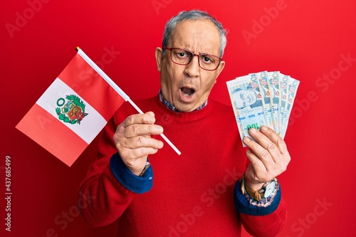Handsome senior man with grey hair holding peru flag and peruvian sol banknotes in shock face, looking skeptical and sarcastic, surprised with open mouth
