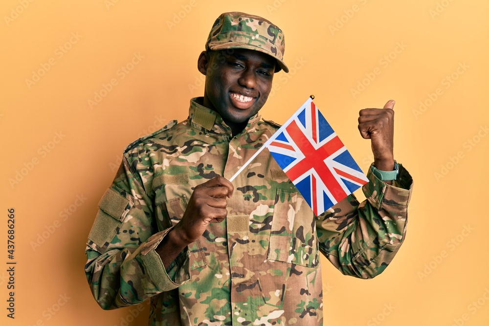Fototapeta premium Young african american man wearing army uniform holding united kingdom flag pointing thumb up to the side smiling happy with open mouth