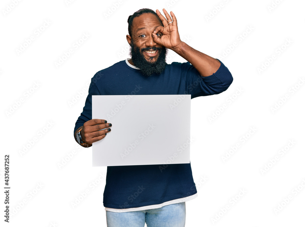 Young african american man holding blank empty banner smiling happy doing ok sign with hand on eye looking through fingers Young african american man holding blank empty banner smiling happy doing ok sign with hand on eye looking through fingers