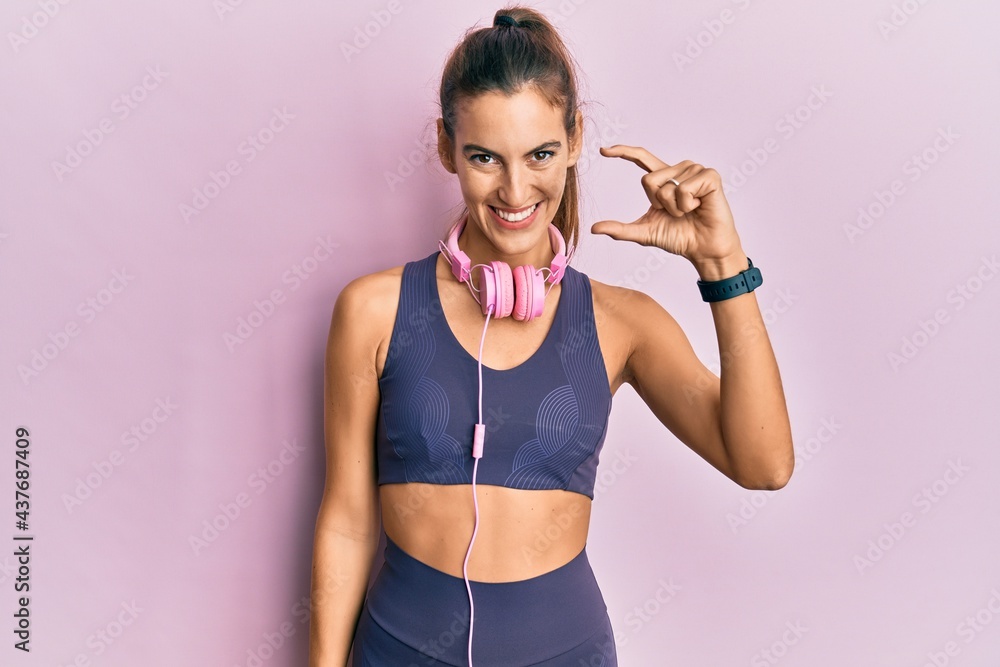 Young beautiful woman wearing gym clothes and using headphones smiling and confident gesturing with hand doing small size sign with fingers looking and the camera. measure concept. Young beautiful woman wearing gym clothes and using headphones smiling and confident gesturing with hand doing small size sign with fingers looking and the camera. measure concept.