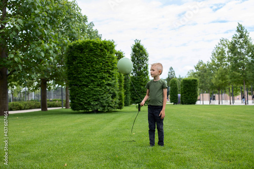 A sad boy in a green T-shirt stands on the grass with a green balloon.