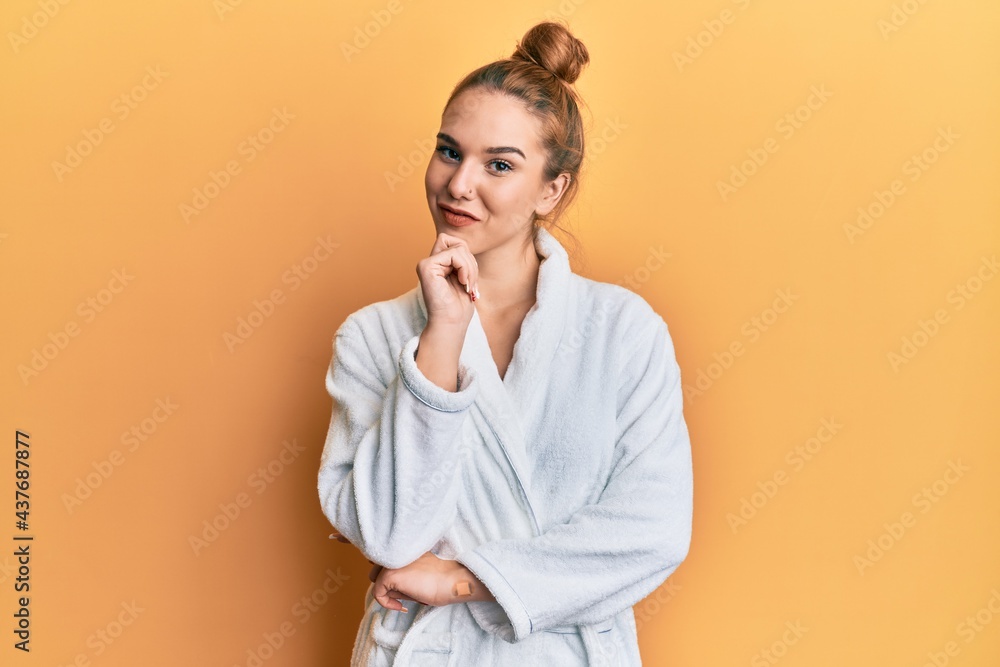 Young blonde woman wearing bathrobe looking confident at the camera with smile with crossed arms and hand raised on chin. thinking positive. Young blonde woman wearing bathrobe looking confident at the camera with smile with crossed arms and hand raised on chin. thinking positive.