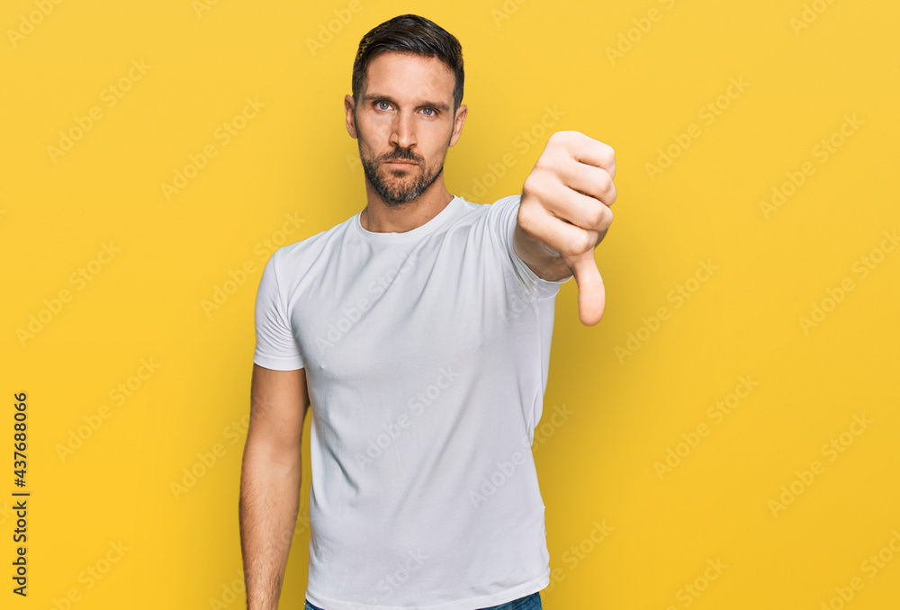 Handsome man with beard wearing casual white t shirt looking unhappy and angry showing rejection and negative with thumbs down gesture. bad expression. Handsome man with beard wearing casual white t shirt looking unhappy and angry showing rejection and negative with thumbs down gesture. bad expression.