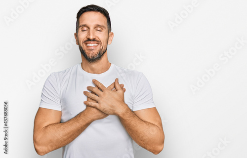 Handsome man with beard wearing casual white t shirt smiling with hands on chest with closed eyes and grateful gesture on face. health concept.
