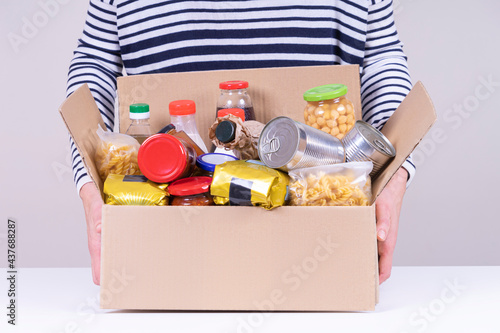 Volunteer female hands holding food donations box with grocery products on white desk. Front view