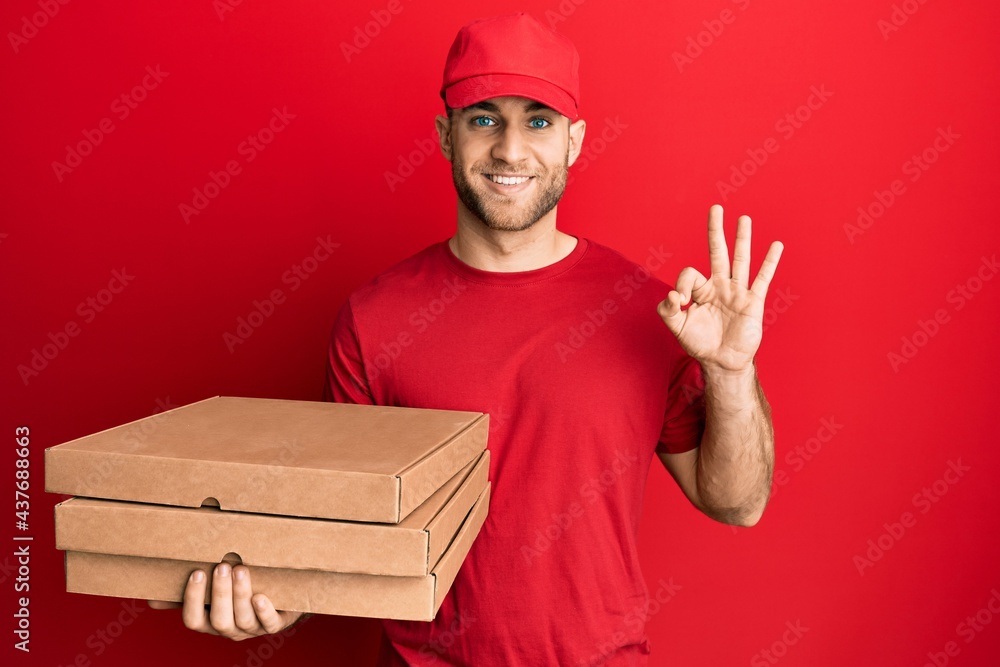 Young caucasian man holding delivery pizza box doing ok sign with fingers, smiling friendly gesturing excellent symbol Young caucasian man holding delivery pizza box doing ok sign with fingers, smiling friendly gesturing excellent symbol