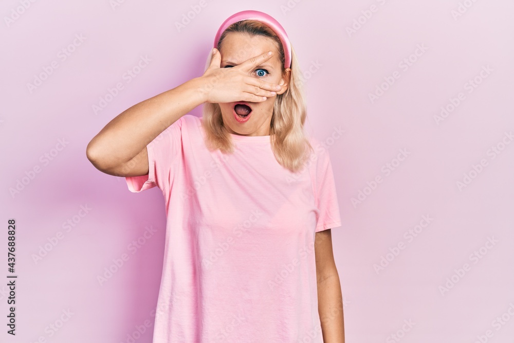 Beautiful caucasian blonde woman wearing casual pink t shirt peeking in shock covering face and eyes with hand, looking through fingers afraid Beautiful caucasian blonde woman wearing casual pink t shirt peeking in shock covering face and eyes with hand, looking through fingers afraid