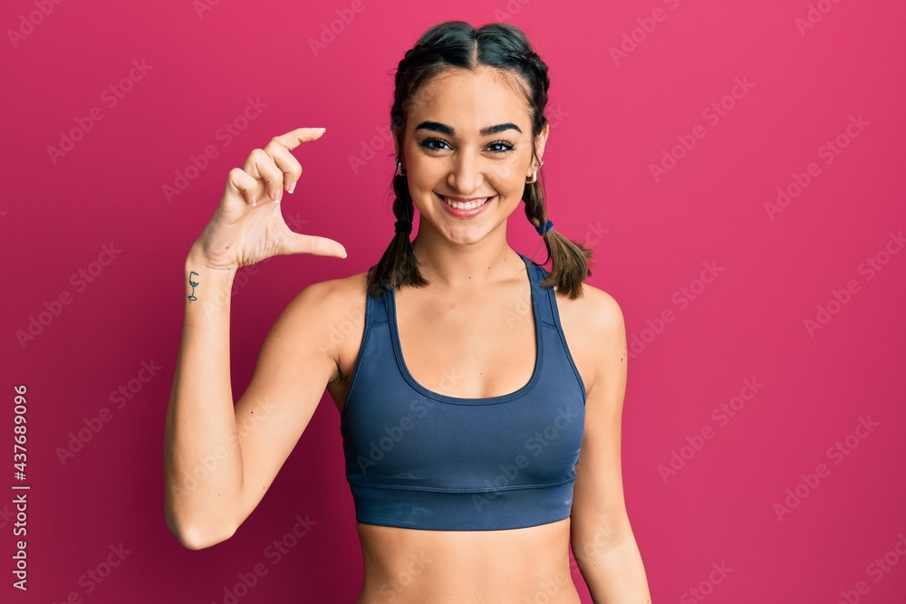 Young brunette girl wearing sportswear and braids smiling and confident gesturing with hand doing small size sign with fingers looking and the camera. measure concept.