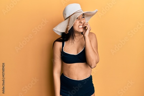 Young latin woman wearing bikini and summer hat looking stressed and nervous with hands on mouth biting nails. anxiety problem.
