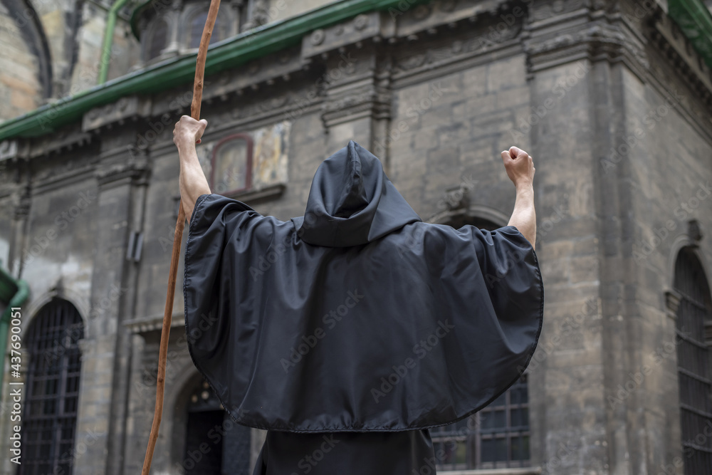 A black-robed monk with a staff stands in front of an old, gloomy building with his staff held high.Concept: curse and mysticism, witchcraft and magic.