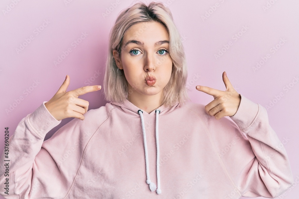 Young blonde girl pointing mouth with fingers puffing cheeks with funny face. mouth inflated with air, catching air. Young blonde girl pointing mouth with fingers puffing cheeks with funny face. mouth inflated with air, catching air.