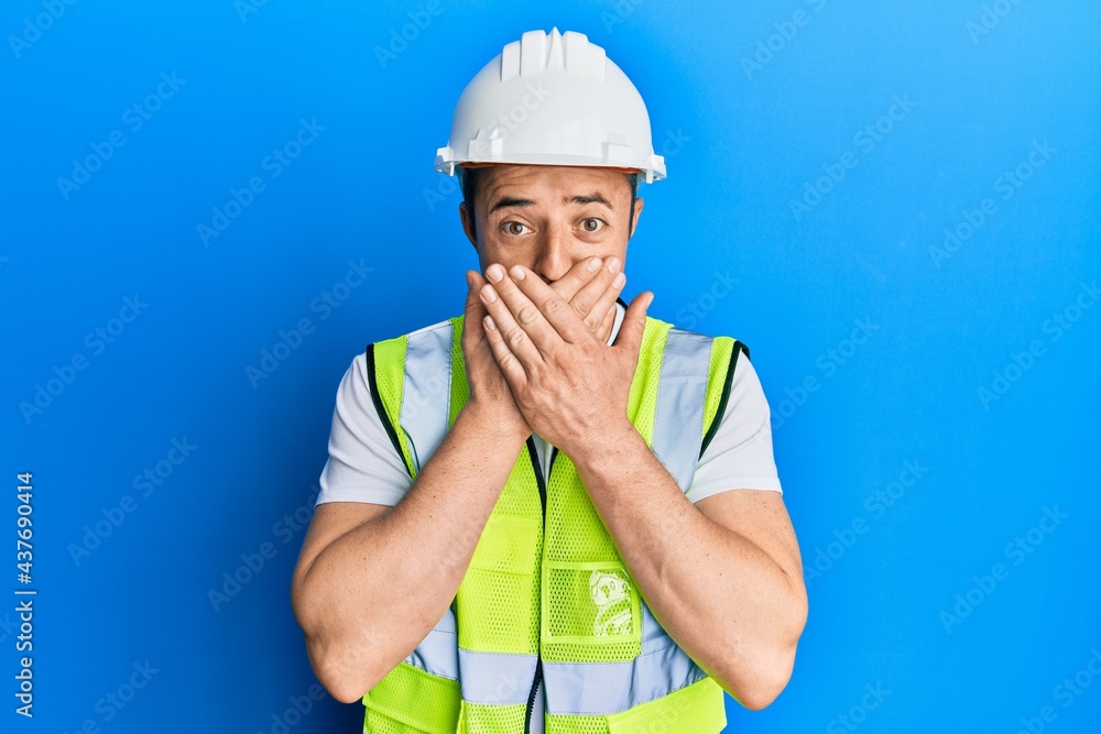 Handsome young man wearing safety helmet and reflective jacket shocked covering mouth with hands for mistake. secret concept.