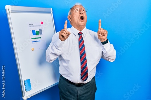 Senior man with grey hair standing by business blackboard amazed and surprised looking up and pointing with fingers and raised arms.