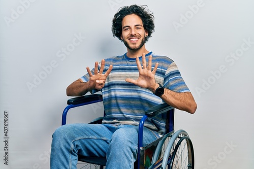 Handsome hispanic man sitting on wheelchair showing and pointing up with fingers number nine while smiling confident and happy.