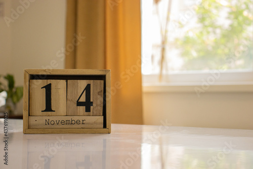 Wooden blocks of the calendar represents the date 14 and the month of November on the background of a window, curtain and a plant.