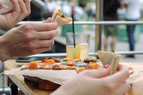 Close-up of hands and pizza with basil leaves in an outdoor street cafe. People eat pizza with their hands. There is lemonade in a glass on the table.