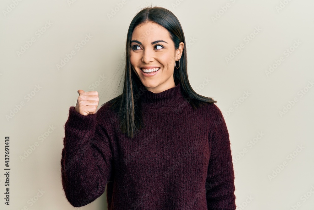 Young brunette woman wearing casual winter sweater pointing thumb up to the side smiling happy with open mouth Young brunette woman wearing casual winter sweater pointing thumb up to the side smiling happy with open mouth