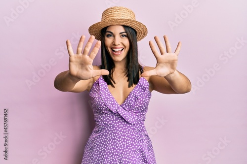 Young brunette woman wearing summer dress and hat showing and pointing up with fingers number ten while smiling confident and happy.