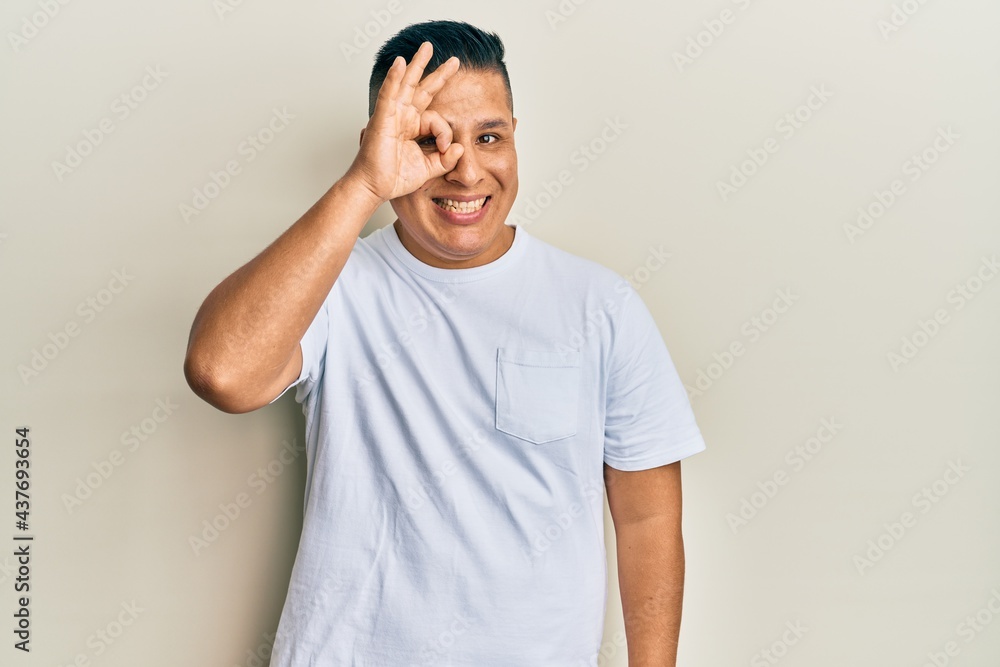Fototapeta premium Young latin man wearing casual white t shirt doing ok gesture with hand smiling, eye looking through fingers with happy face.