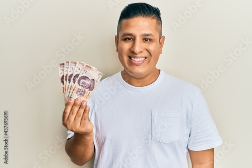 Young latin man holding 500 mexican pesos banknotes looking positive and happy standing and smiling with a confident smile showing teeth photo