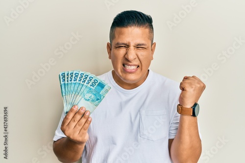 Young latin man holding 100 brazilian real banknotes screaming proud, celebrating victory and success very excited with raised arm