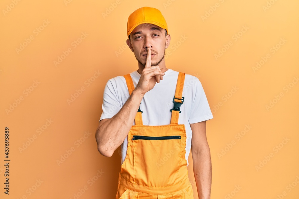 Hispanic young man wearing handyman uniform asking to be quiet with finger on lips. silence and secret concept. Hispanic young man wearing handyman uniform asking to be quiet with finger on lips. silence and secret concept.