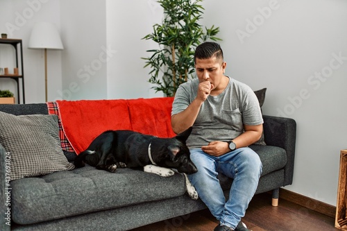 Young latin man and dog sitting on the sofa at home feeling unwell and coughing as symptom for cold or bronchitis. health care concept.