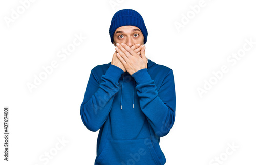 Hispanic young man wearing sweatshirt and wool hat shocked covering mouth with hands for mistake. secret concept.