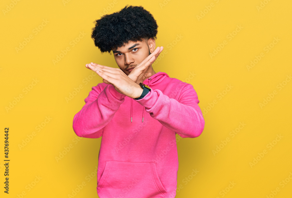 Young african american man with afro hair wearing casual pink sweatshirt rejection expression crossing arms and palms doing negative sign, angry face Young african american man with afro hair wearing casual pink sweatshirt rejection expression crossing arms and palms doing negative sign, angry face
