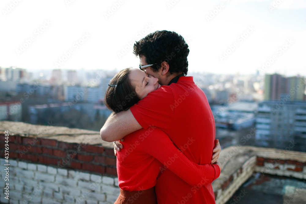 Man and caucasian woman hug each other tightly and kiss on the roof of a tall building overlooking the city. Lovers love each other and both are dressed in red. Man and caucasian woman hug each other tightly and kiss on the roof of a tall building overlooking the city. Lovers love each other and both are dressed in red.