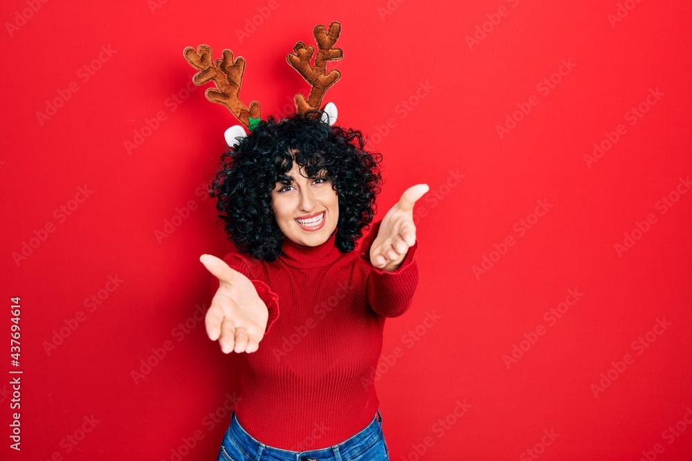 Young middle east woman wearing cute christmas reindeer horns looking at the camera smiling with open arms for hug. cheerful expression embracing happiness. Young middle east woman wearing cute christmas reindeer horns looking at the camera smiling with open arms for hug. cheerful expression embracing happiness.