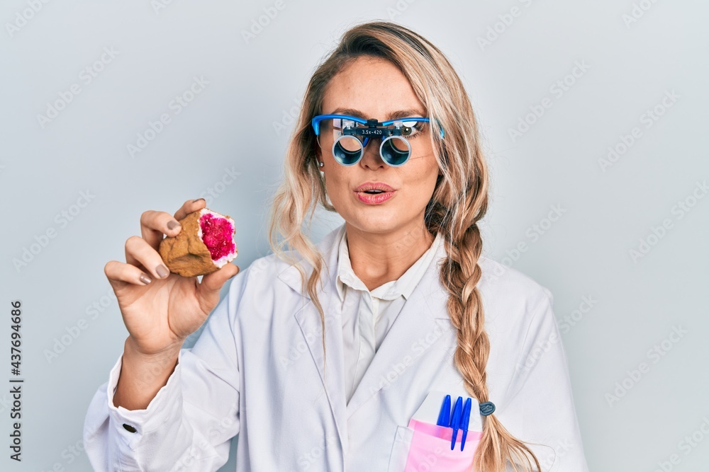 Obraz premium Beautiful young blonde woman holding geode stone wearing magnifier glasses scared and amazed with open mouth for surprise, disbelief face