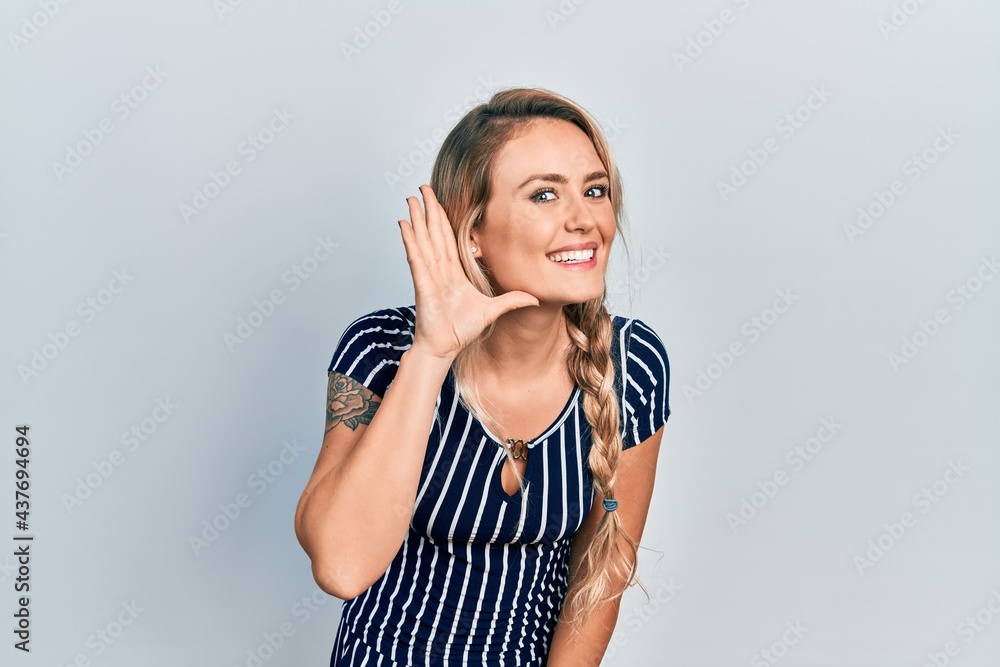 Beautiful young blonde woman wearing casual striped dress smiling with hand over ear listening an hearing to rumor or gossip. deafness concept. Beautiful young blonde woman wearing casual striped dress smiling with hand over ear listening an hearing to rumor or gossip. deafness concept.