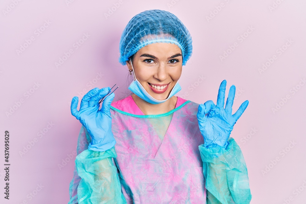 Young brunette woman wearing surgeon uniform holding tweezers doing ok sign with fingers, smiling friendly gesturing excellent symbol Young brunette woman wearing surgeon uniform holding tweezers doing ok sign with fingers, smiling friendly gesturing excellent symbol