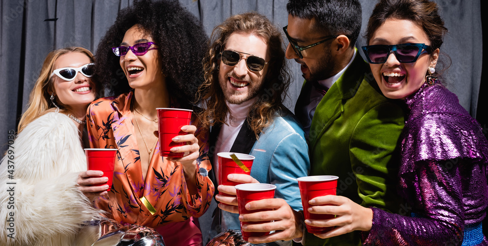 playful interracial friends in sunglasses celebrating with plastic cups on grey background, banner.