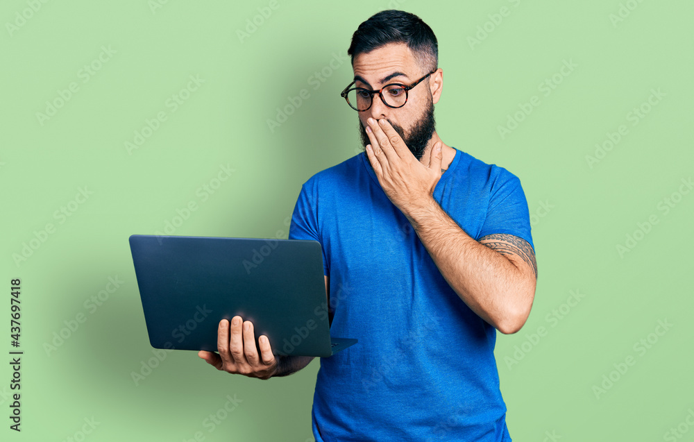 Hispanic man with beard working using computer laptop covering mouth with hand, shocked and afraid for mistake. surprised expression Hispanic man with beard working using computer laptop covering mouth with hand, shocked and afraid for mistake. surprised expression