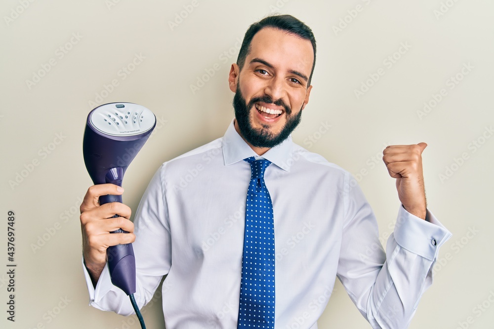 Young business man with beard holding electric steam iron pointing thumb up to the side smiling happy with open mouth Young business man with beard holding electric steam iron pointing thumb up to the side smiling happy with open mouth
