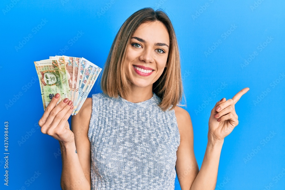 Young caucasian blonde woman holding peruvian sol banknotes smiling happy pointing with hand and finger to the side - obrazy, fototapety, plakaty 
