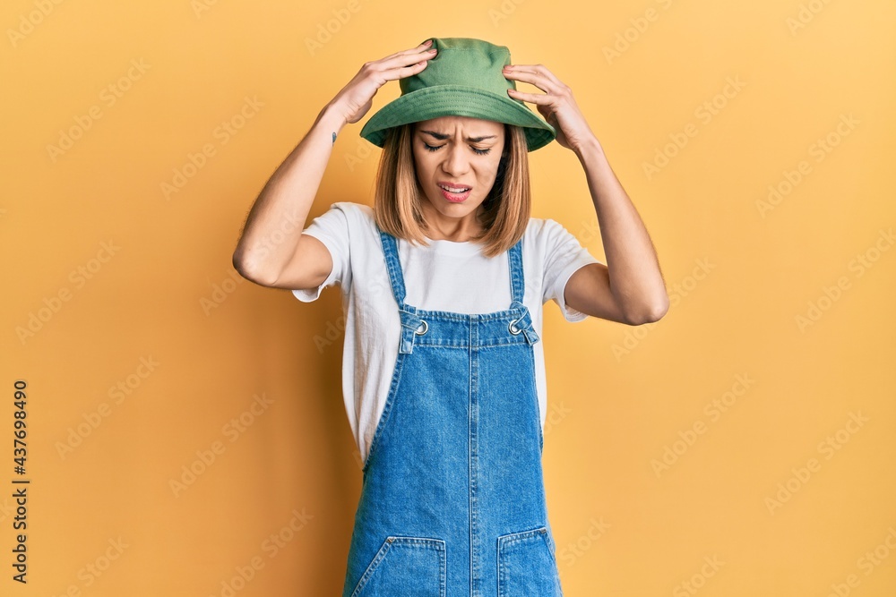 Young caucasian blonde woman wearing denim jumpsuit and hat with 90s style suffering from headache desperate and stressed because pain and migraine. hands on head. Young caucasian blonde woman wearing denim jumpsuit and hat with 90s style suffering from headache desperate and stressed because pain and migraine. hands on head.