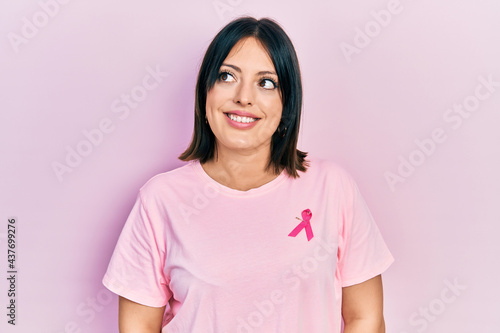 Young hispanic woman wearing pink cancer ribbon on t shirt smiling looking to the side and staring away thinking.