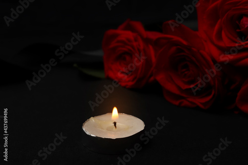 Burning mourning candle and red roses with black mourning ribbon on a dark background. The concept of grief, death, remembrance of the