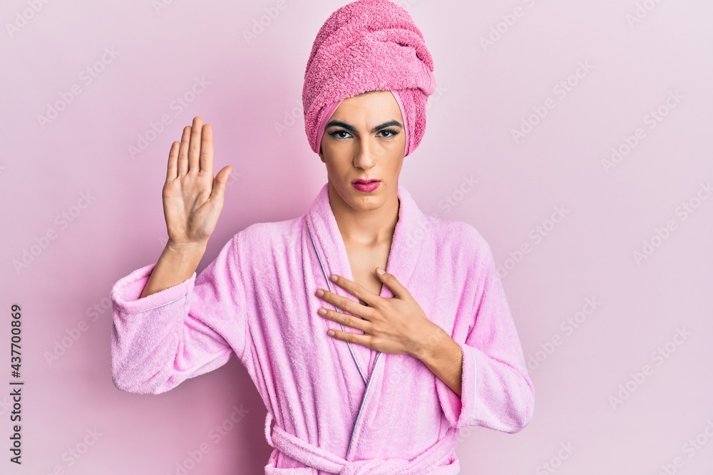 Young man wearing woman make up wearing shower towel on head and bathrobe swearing with hand on chest and open palm, making a loyalty promise oath Young man wearing woman make up wearing shower towel on head and bathrobe swearing with hand on chest and open palm, making a loyalty promise oath