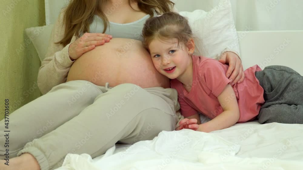 girl lies in bed with pregnant woman. Daughter hugs mom's belly. Pregnant mother with second child and older sister happy together at home. preschool girl listens with ear of unborn baby inside tummy